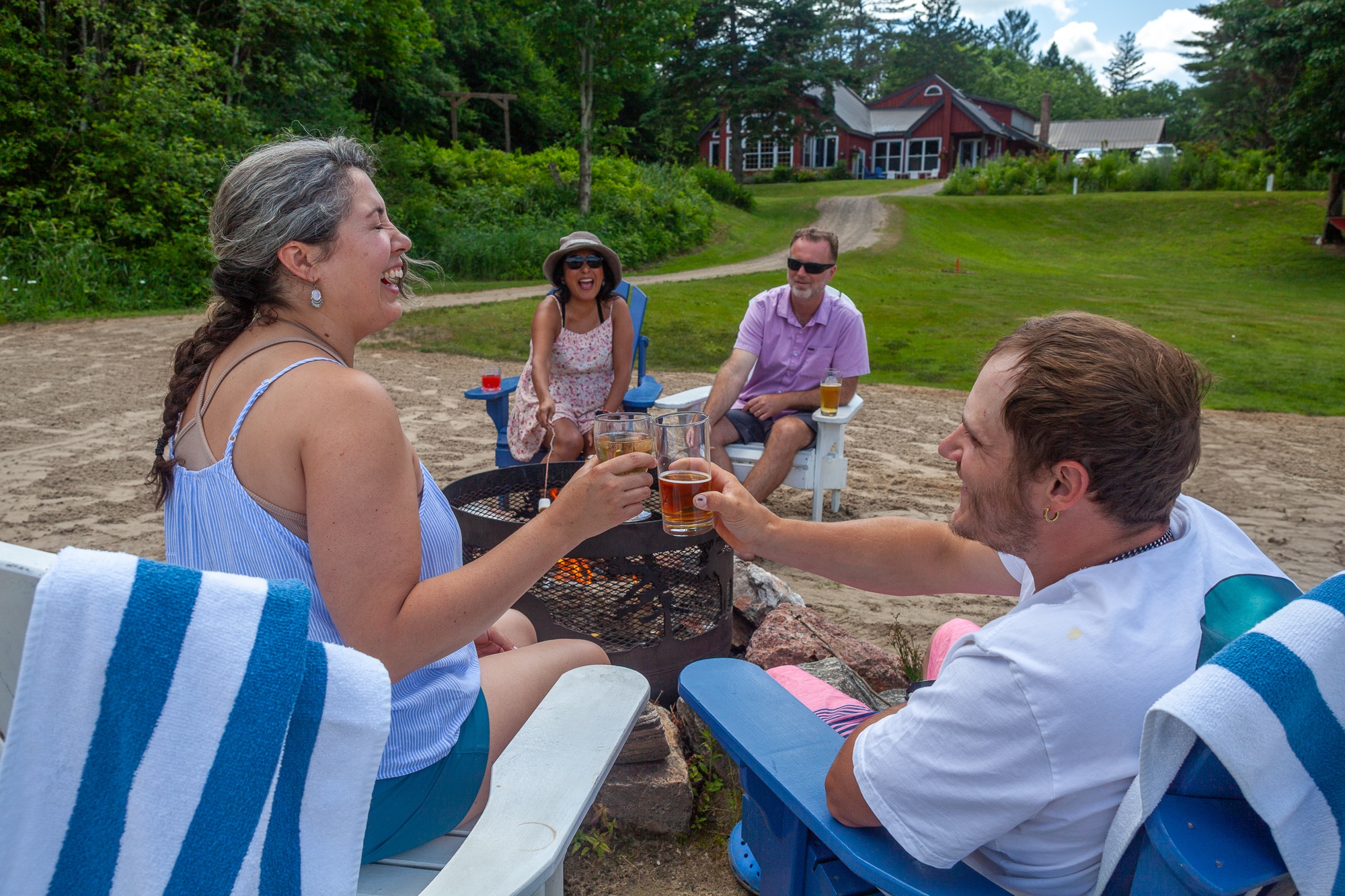 Guests toasting drinks around beachside fire pit at Trillium Resort and Spa with lodge and lake in summer Muskoka Huntsville Port Sydney
